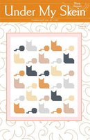 Pattern cover for 'Under My Skein' quilt by Wendy Sheppard, featuring a white quilt design with rows of stylized cat silhouettes in orange, beige, gray, and dark charcoal, framed by a pink border on an orange and white patterned background.